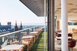 Modern rooftop café terrace with rows of wooden tables and cushioned chairs on green turf behind a glass railing overlooking a city skyline with church spires, and an adjacent indoor dining space featuring round tables and red leather armchairs under circular ceiling panels.