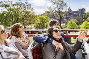 A group of cheerful tourists enjoying the view from the upper deck of an open-top tour bus in Edinburgh, surrounded by greenery and historic architecture.