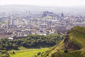 Panoramic view of Edinburgh: Edinburgh Castle perched on Castle Rock with the city’s historic skyline behind, and the green grassy slopes of Holyrood Park in the foreground.