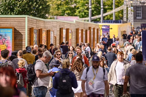 Large crowd of people walking through an outdoor festival or market area during the day, with posters displayed on wooden booths and trees in the background.