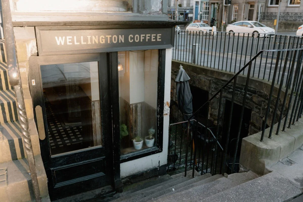 Wellington Coffee, a small and cozy café located in a basement-level space with a black-framed entrance and large window. The café's name is displayed above the door in white lettering. A descending staircase with iron railings leads to an outdoor seating area, partially hidden in shadow. The surrounding urban environment includes parked cars and stone buildings.
