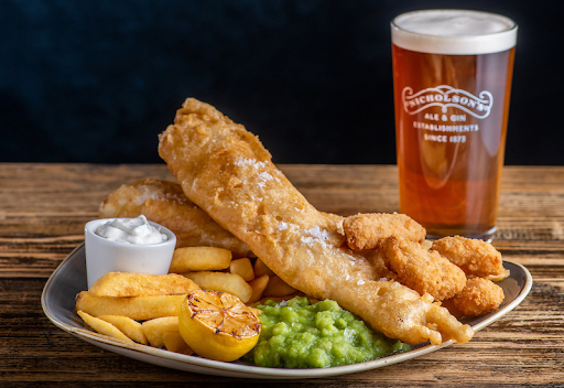 Classic British fish and chips with mushy peas, tartar sauce, and a pint of Nicholson’s beer on a rustic wooden table.
