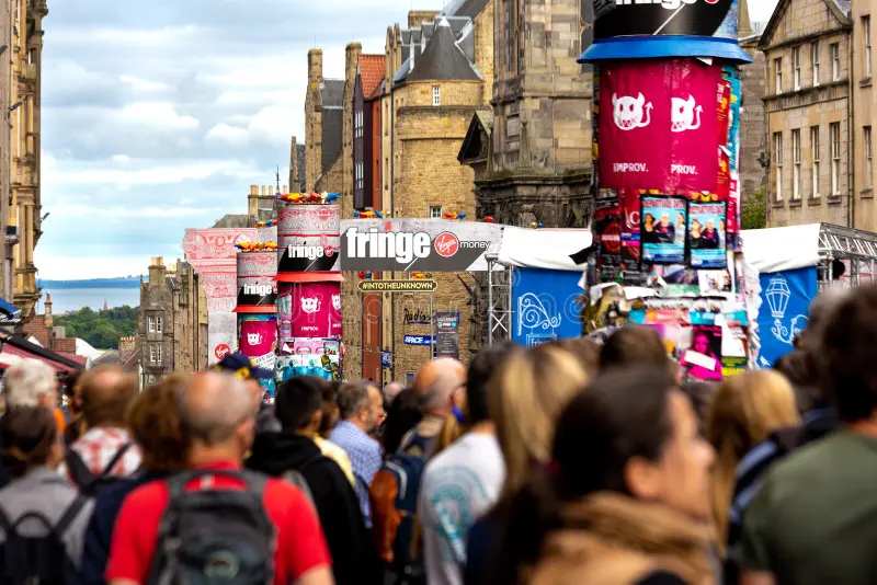 A bustling scene at the Edinburgh Festival Fringe, with crowds filling the historic street and brightly colored posters advertising shows on tall pillars.
