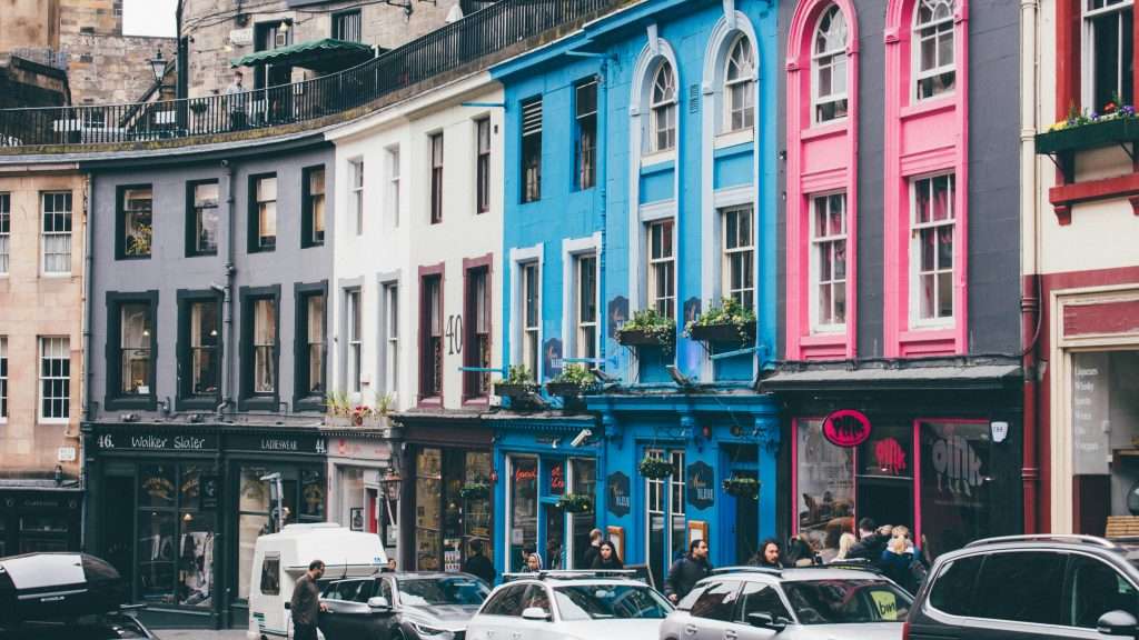 A lively street in Edinburgh showcasing colorful, historic storefronts and bustling foot traffic, with bright facades in shades of blue and pink drawing attention along the curved row of buildings.
