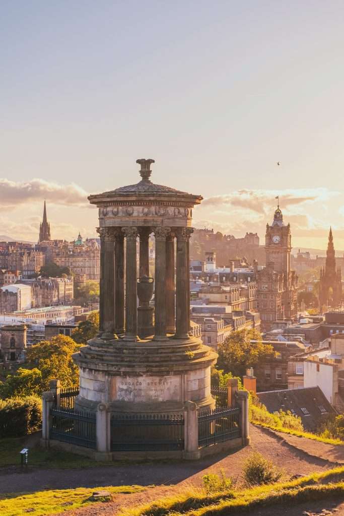 A historic neoclassical monument perched on Calton Hill overlooking Edinburgh’s skyline, with a prominent clock tower and Gothic spires bathed in golden evening light.