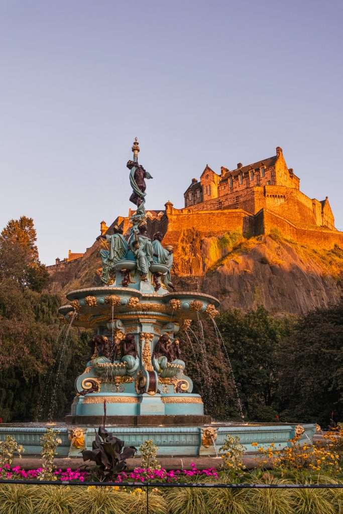 An ornate fountain surrounded by colorful flowers, with the dramatic silhouette of Edinburgh Castle perched high on the rocky crag behind it, lit by the warm glow of the evening sun.