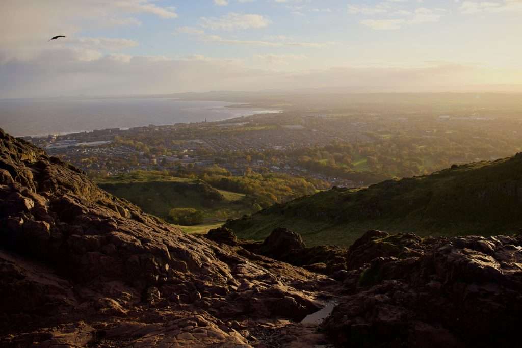 A sweeping panoramic view of Edinburgh and its coastline from a rugged hillside, with rolling greenery and a golden evening light illuminating the city below.