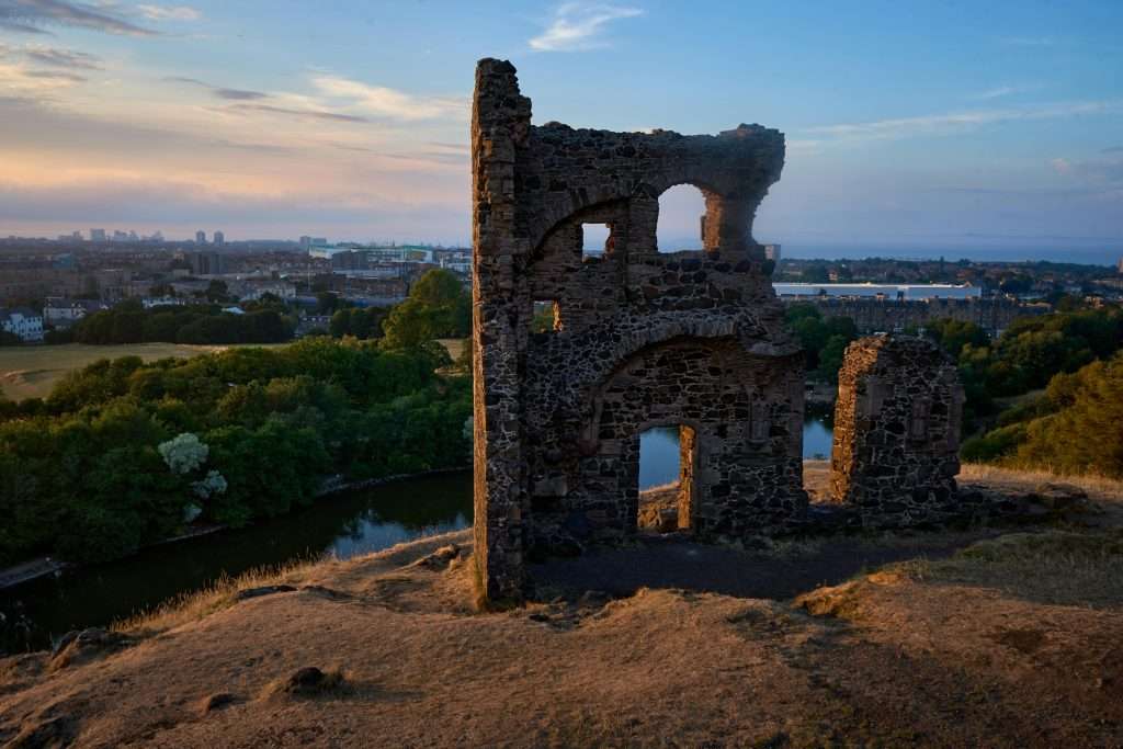 The partial remains of an ancient stone structure perched on a hillside in Holyrood Park, overlooking Edinburgh’s urban landscape and lush greenery at sunset.