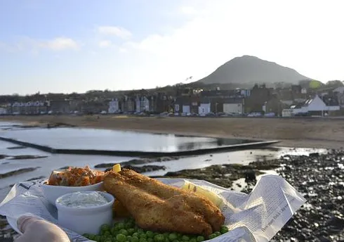 Golden battered fish fillets with peas, lemon wedge, tartar sauce and orange relish, seaside view.
