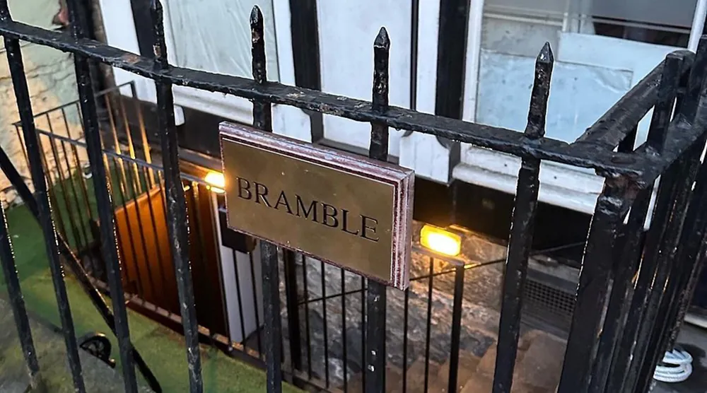 Brass Bramble sign on black railing above stone stairwell to basement bar entrance.