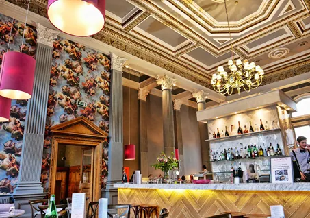 Elegant bar with marble counter, chandelier lighting, and classical wall art in a grand interior.