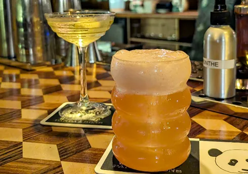 Frothy cocktail in sculpted glass and sparkling drink in coupe on panda-themed coasters.