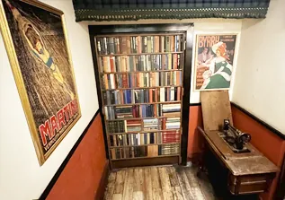 Bookshelf door in vintage hallway with antique sewing machine and retro posters on walls.