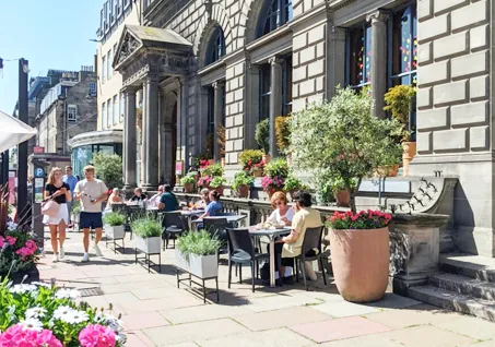 Outdoor café with people dining, surrounded by potted plants, flowers, and a historic stone building.