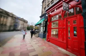 Red-painted bar storefront with ornate woodwork, pedestrians walking on a stone-paved street.