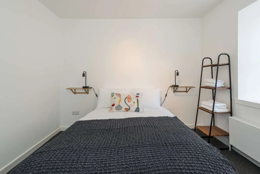 Bedroom with double bed, dark blanket, patterned pillows, and shelving unit with folded white towels