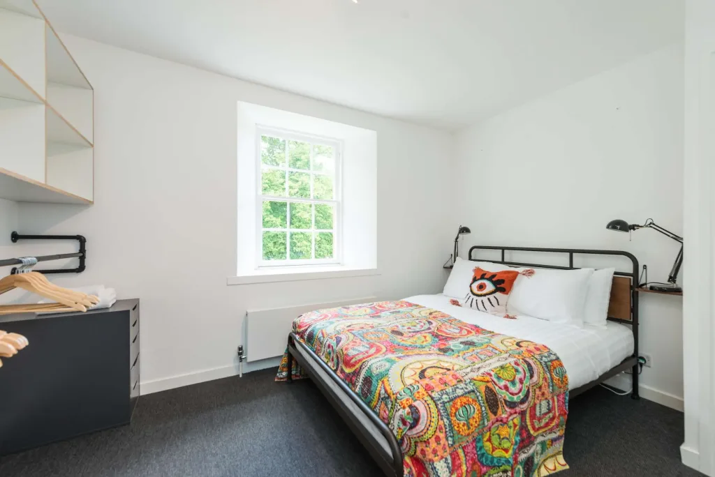 Bright modern bedroom with double bed, colorful patterned quilt, quirky eye pillow, reading lamps, and large window view