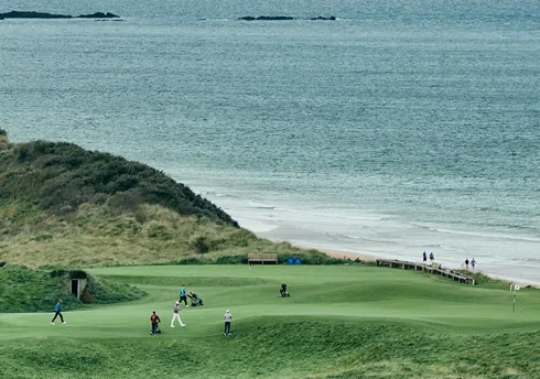Coastal links golf green with golfers beside sandy beach and sea.