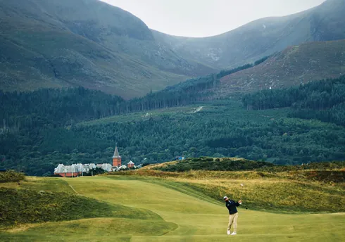 Golfer swinging on mountain valley fairway with forested hills and village buildings.