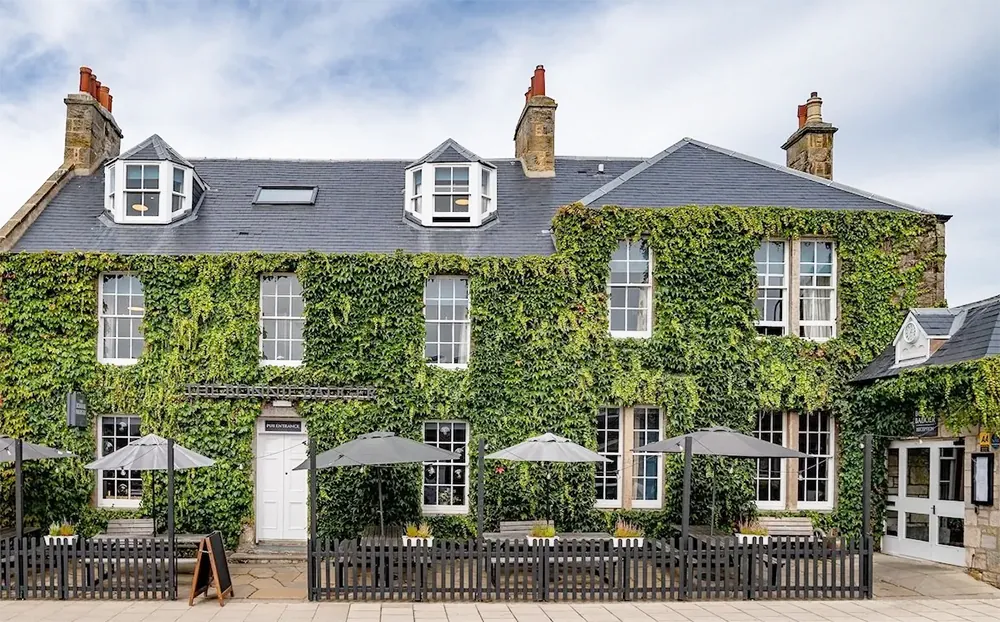 Ivy-covered inn facade with dormer windows, outdoor seating and umbrellas.