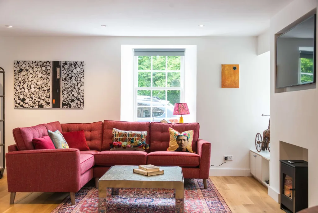 Living room with red corner sofa, patterned cushions, coffee table with books, wall art, and flat-screen TV above a fireplace