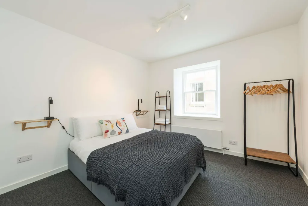 Minimalist bedroom with double bed, grey blanket, decorative pillows, open clothes rack and window light