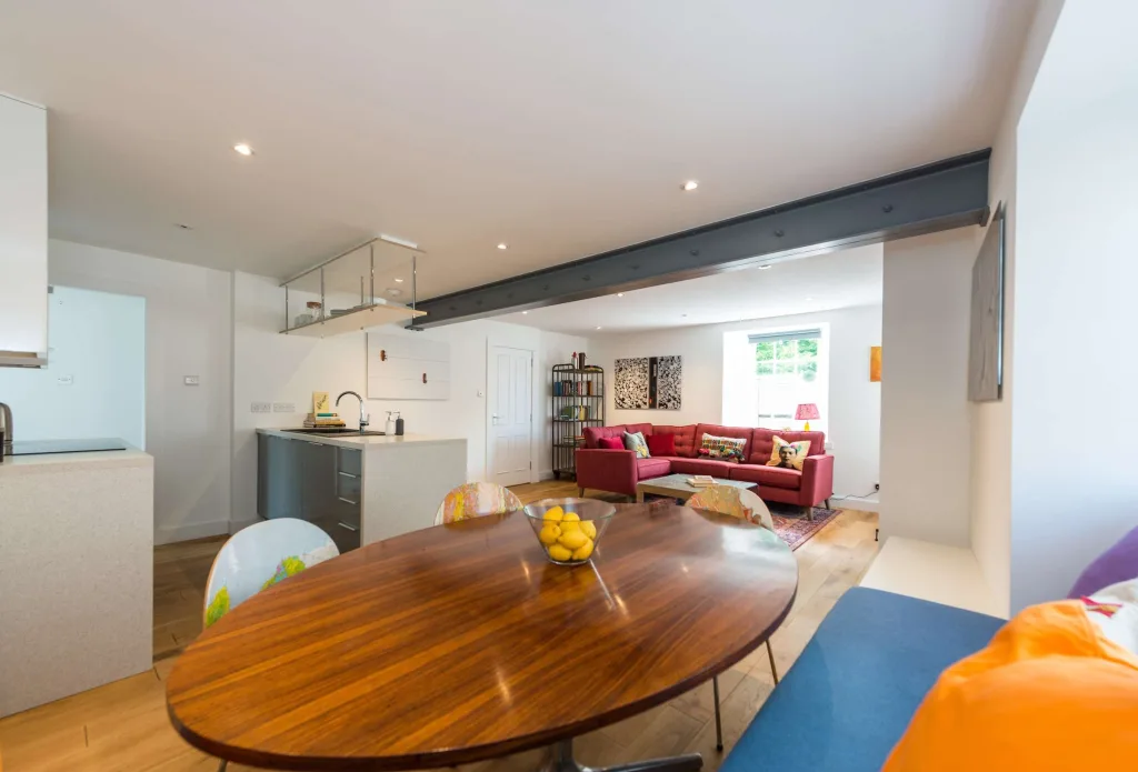 Open-plan living room with modern kitchen, wooden dining table, and red corner sofa in holiday cottage