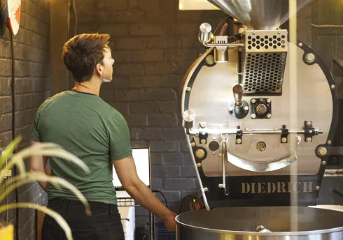 Person operating Diedrich coffee roaster in warm, industrial brick roastery.