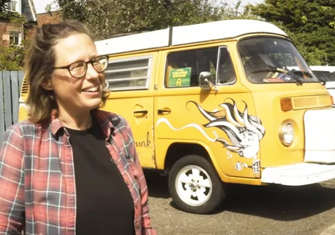 Smiling woman beside yellow vintage camper van with mural, sunny outdoor setting.