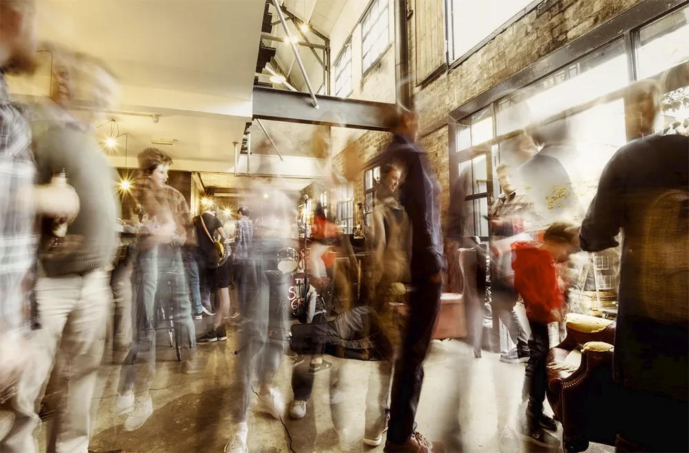 Busy industrial interior with blurred people, brick walls, large windows, warm ambient lighting.