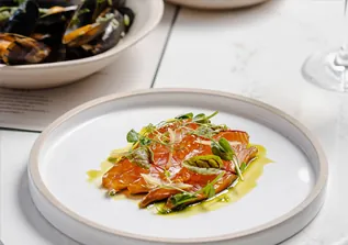 Thinly sliced fish with microgreens and oil on white plate, mussels nearby.