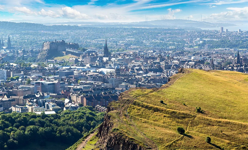 View of city skyline with castle, church spires, and grassy hill in foreground.