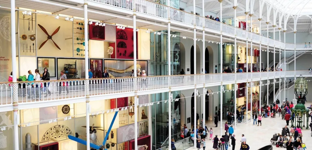 Crowds exploring multi-level museum hall with glass displays, artifacts, and ornate white balconies.