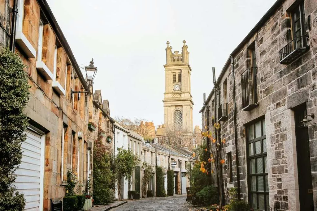 Stone-paved lane with stone houses, greenery, and a tall clock tower.